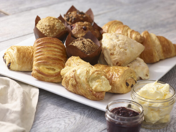 Assorted pastries on a square white platter with croissants, muffins and danish with a mason jars of butter and jam on a grey wooden background