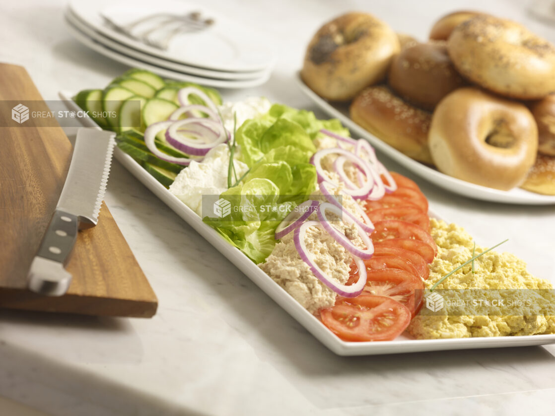 White rectangular platter with tuna salad, egg salad, cream cheese, sliced cucumber, red onion, tomatoes with whole bagels in the background on a marble background