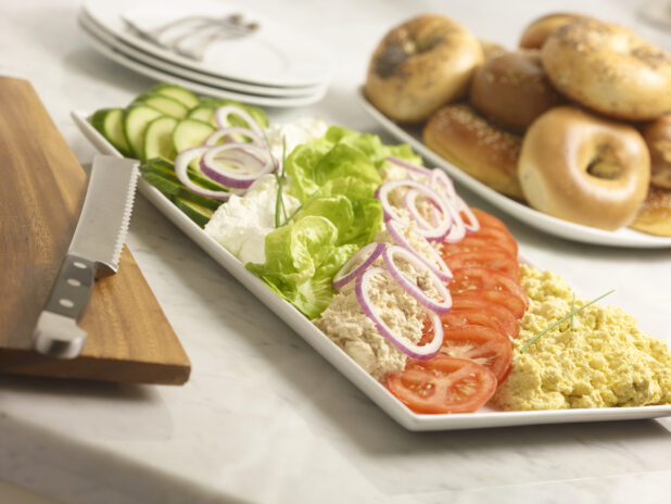 White rectangular platter with tuna salad, egg salad, cream cheese, sliced cucumber, red onion, tomatoes with whole bagels in the background on a marble background