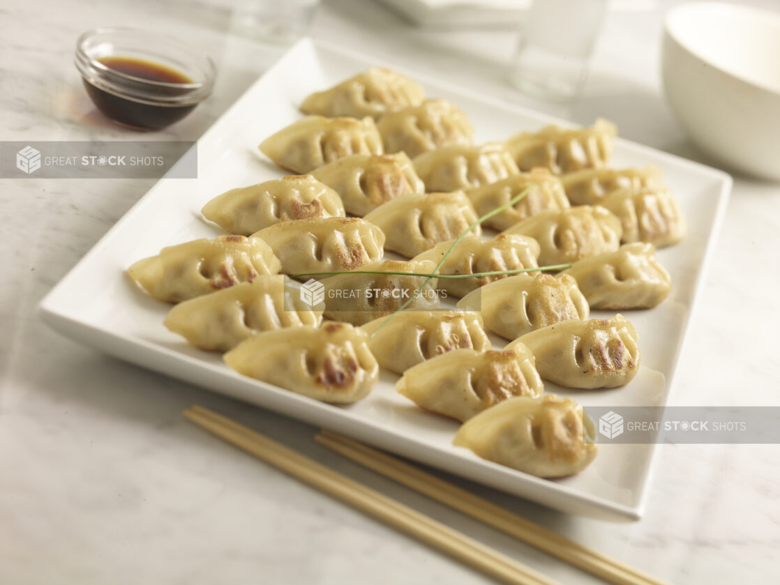 Chinese pan fried dumplings on a white square platter with soya sauce in the background and chopsticks in the foreground on a white marble background