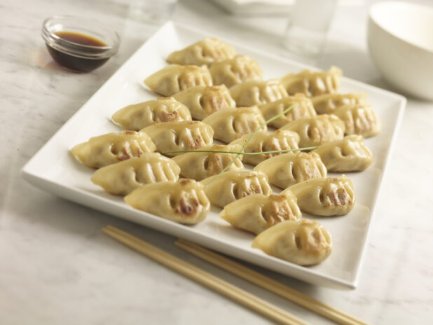 Chinese pan fried dumplings on a white square platter with soya sauce in the background and chopsticks in the foreground on a white marble background