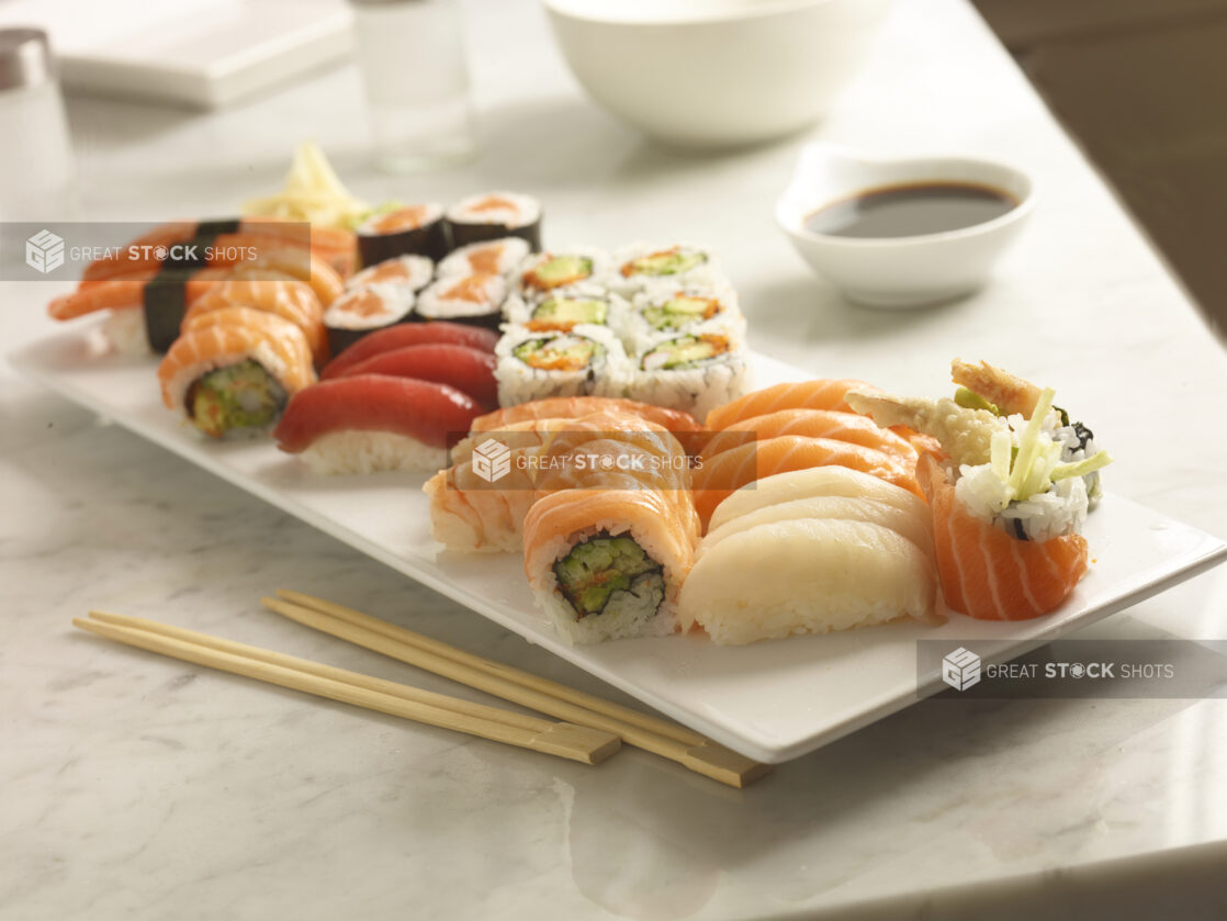 Assorted sushi on a white rectangular platter with chopsticks in the foreground and soya sauce in the background on a white marble background