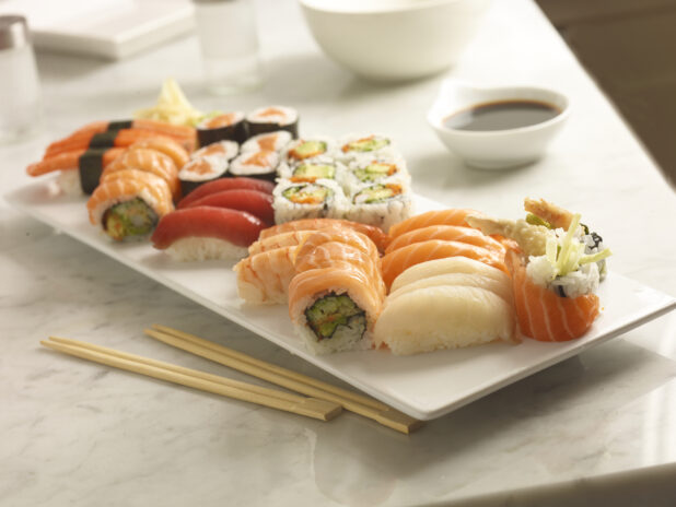 Assorted sushi on a white rectangular platter with chopsticks in the foreground and soya sauce in the background on a white marble background