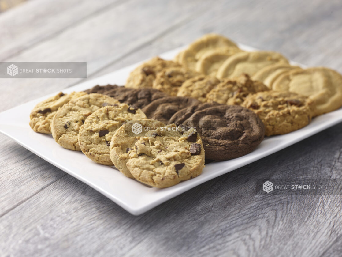 Assorted fresh baked cookies arranged in rows on a white rectangular platter on a grey wooden background