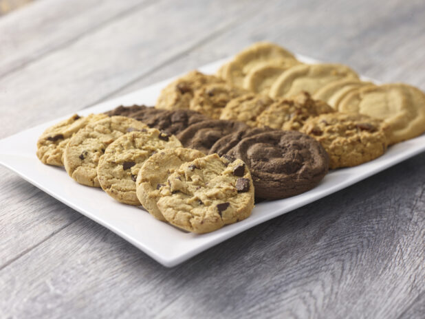 Assorted fresh baked cookies arranged in rows on a white rectangular platter on a grey wooden background