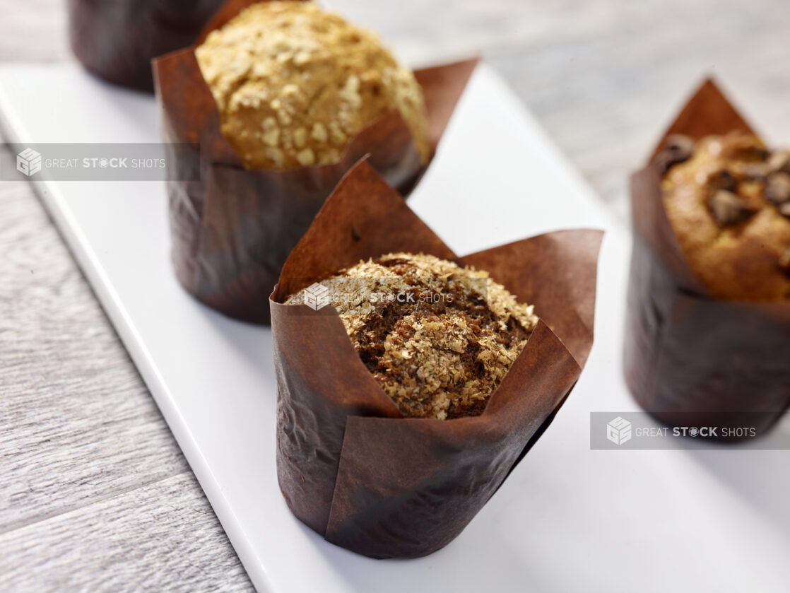 Assorted muffins wrapped in brown parchment paper on a white rectangular board in a close up view