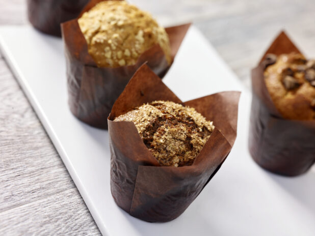 Assorted muffins wrapped in brown parchment paper on a white rectangular board in a close up view