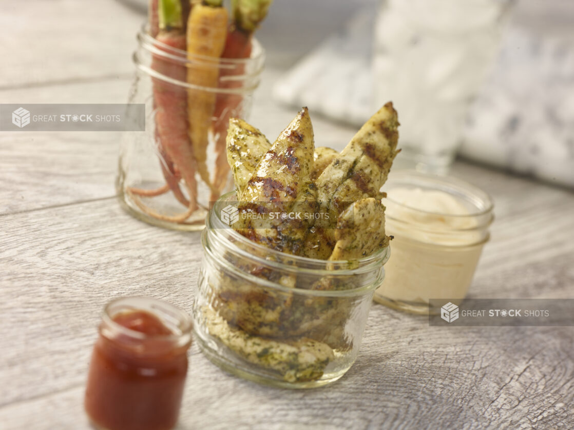 Grilled chicken tenders in a mason jar in the foreground with heirloom carrots, mayo and ketchup in various mason jars in the background