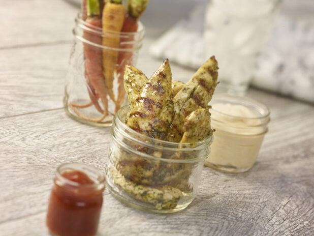 Grilled chicken tenders in a mason jar in the foreground with heirloom carrots, mayo and ketchup in various mason jars in the background