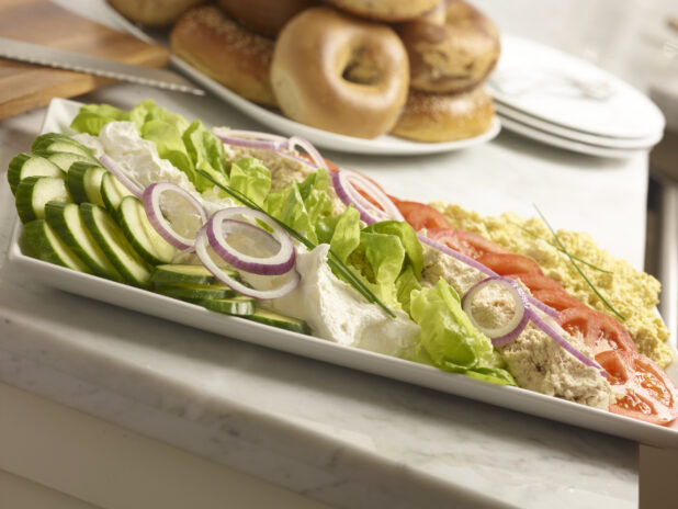 White rectangular platter with tuna salad, egg salad, cream cheese, sliced cucumber, red onion, tomatoes with whole bagels in the background on a marble background