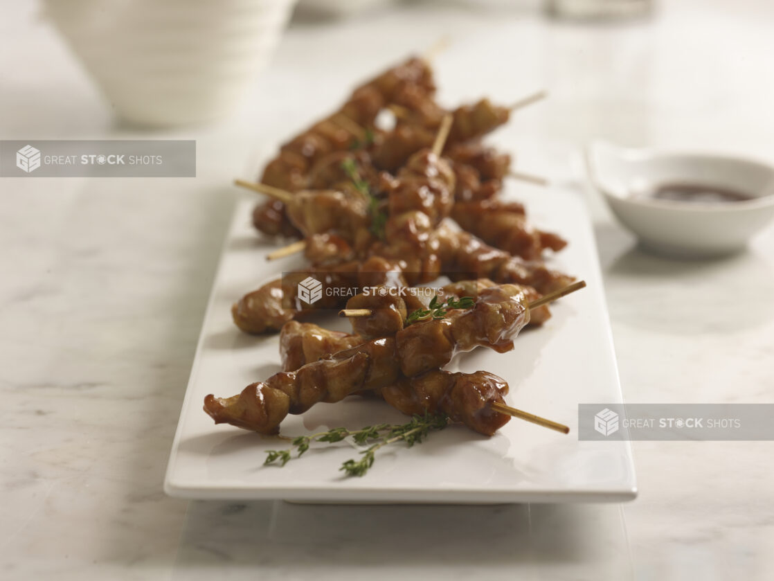 Chicken teriyaki skewers on a white rectangular platter with fresh thyme sprigs and dipping sauce in a small white bowl in the background