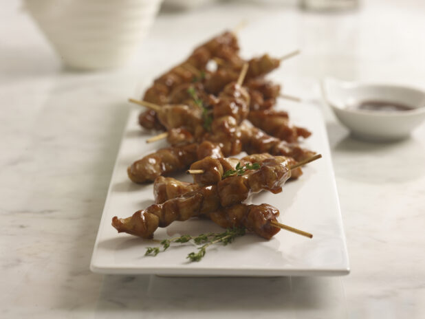 Chicken teriyaki skewers on a white rectangular platter with fresh thyme sprigs and dipping sauce in a small white bowl in the background