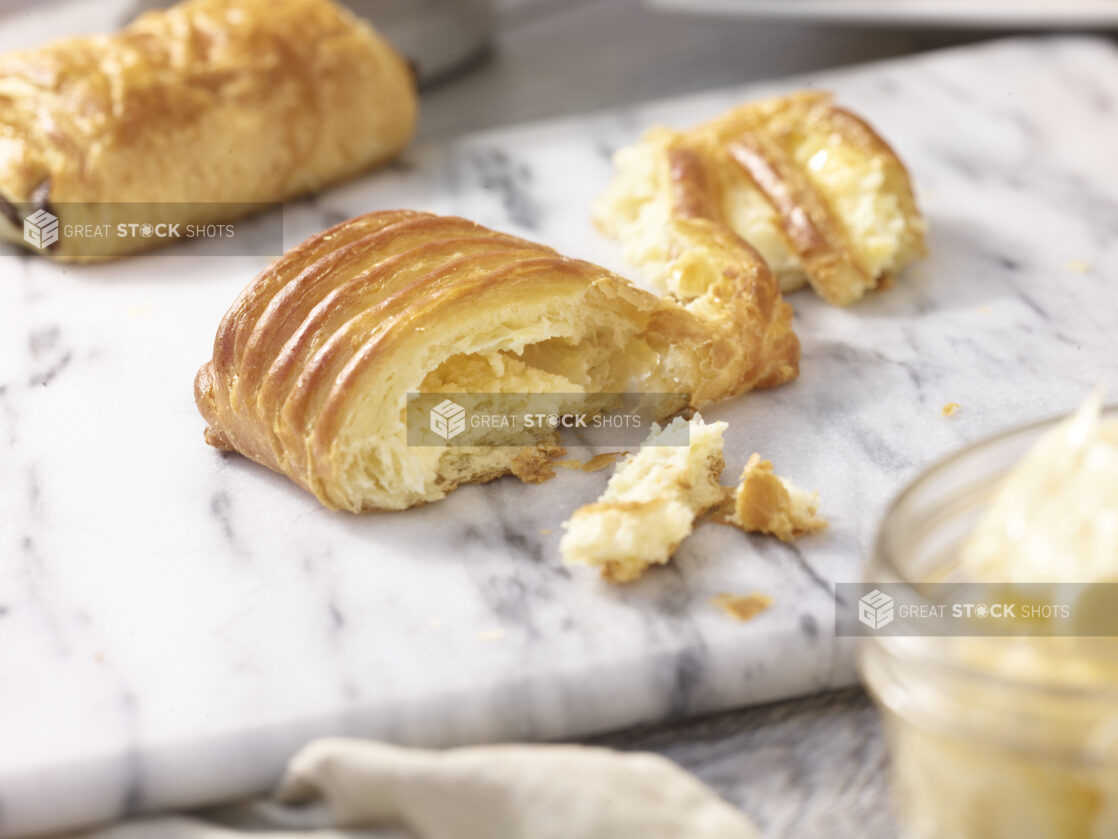 A danish torn in half on a white marble board with a chocolate danish in the background and a jar of butter in the foreground