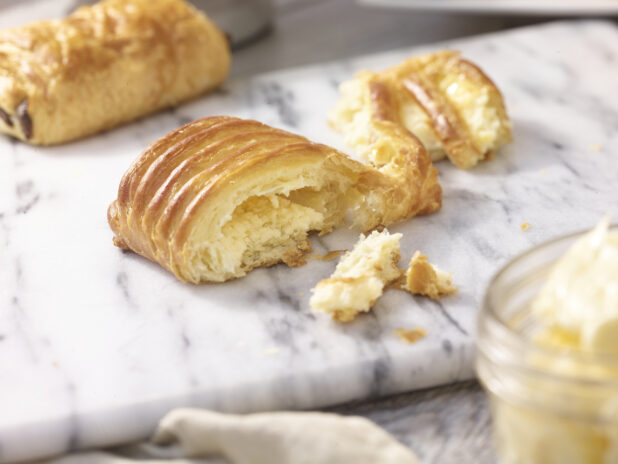 A danish torn in half on a white marble board with a chocolate danish in the background and a jar of butter in the foreground