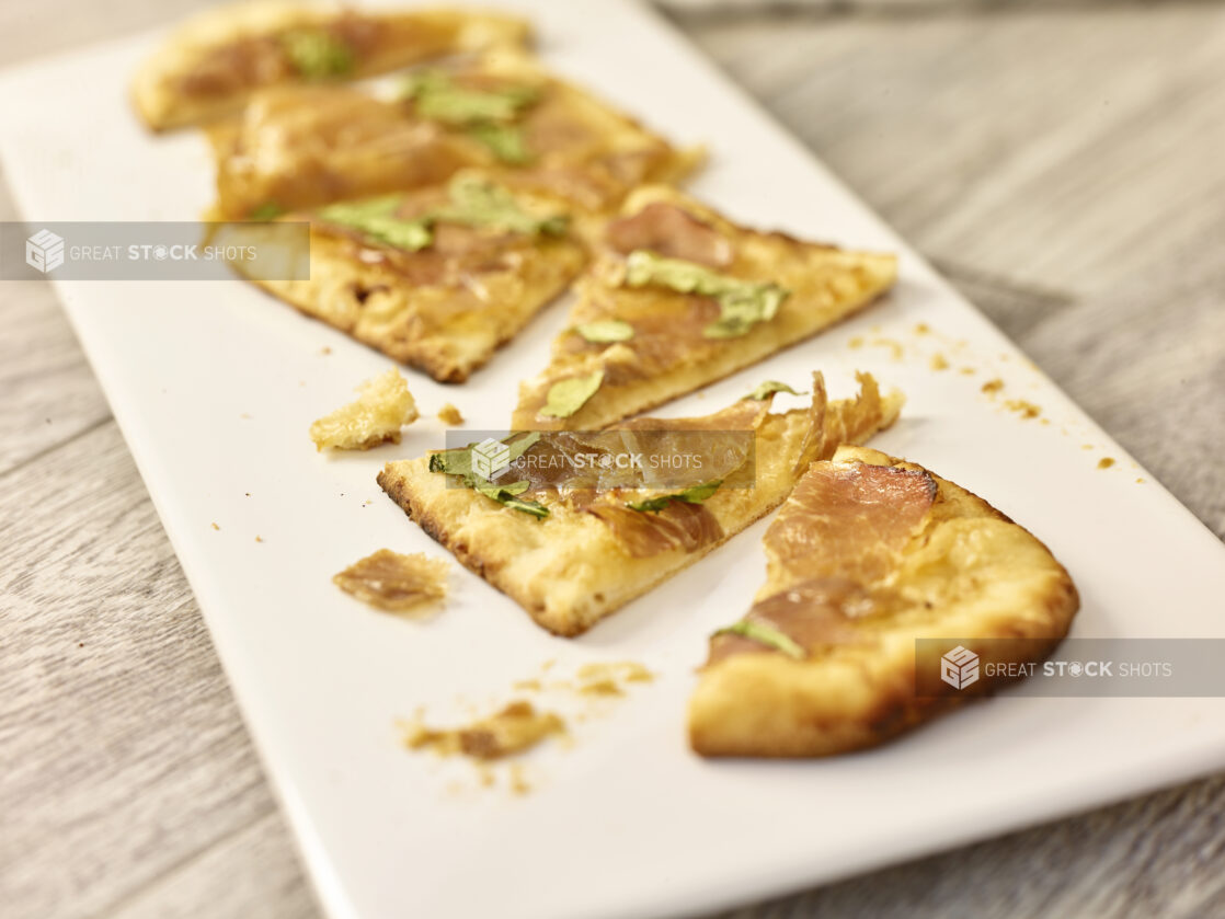 Slices of prosciutto and arugula flatbread on a white rectangular platter on a grey wooden background