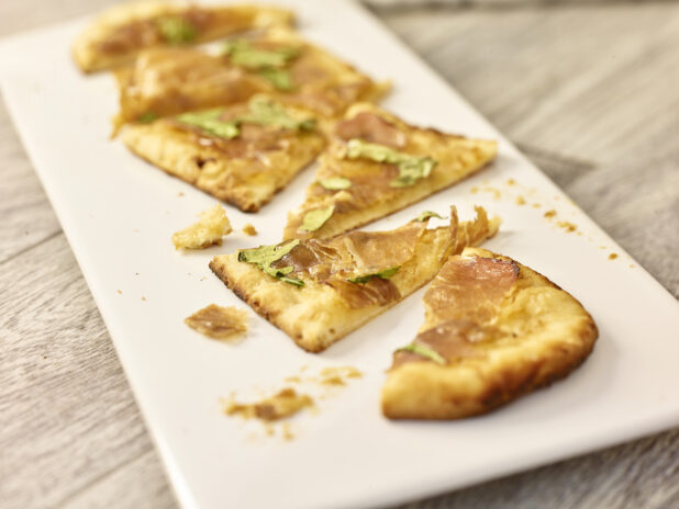 Slices of prosciutto and arugula flatbread on a white rectangular platter on a grey wooden background