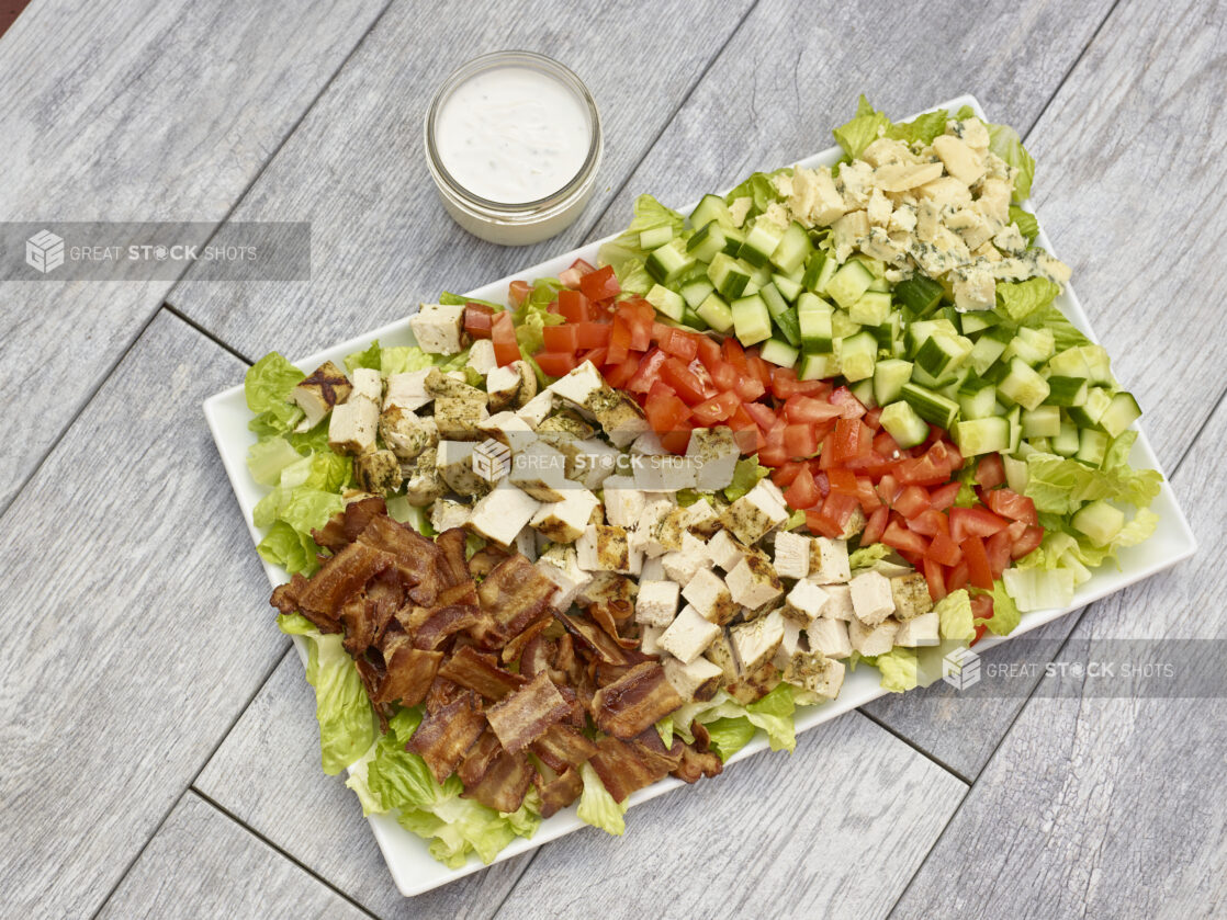 Large platter of chicken BLT salad with grilled chicken, bacon, romaine lettuce, cucumber, tomato and blue cheese with dressing in a mason jar all on a grey wooden background