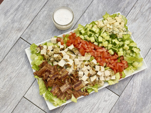 Large platter of chicken BLT salad with grilled chicken, bacon, romaine lettuce, cucumber, tomato and blue cheese with dressing in a mason jar all on a grey wooden background