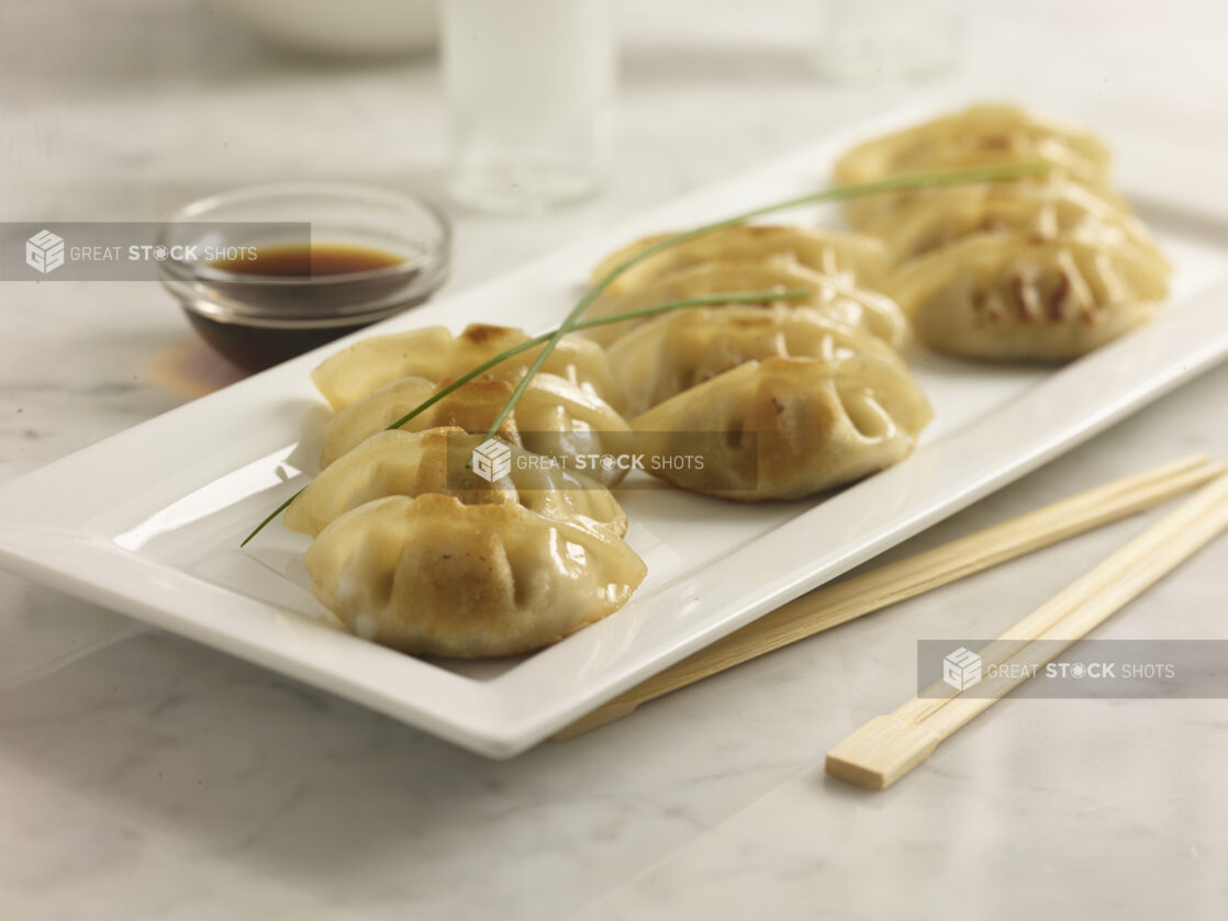 Pan fried Chinese dumplings on a white rectangular platter with soya sauce and chopsticks on a white marble background