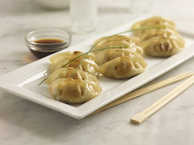 Pan fried Chinese dumplings on a white rectangular platter with soya sauce and chopsticks on a white marble background