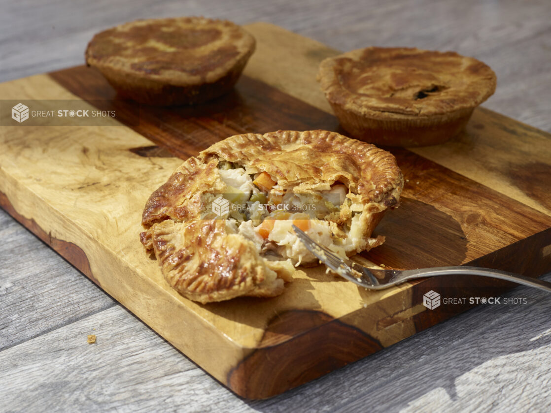 Individual chicken pot pies on a wooden board on a grey wooden background, one pie broken open with a fork