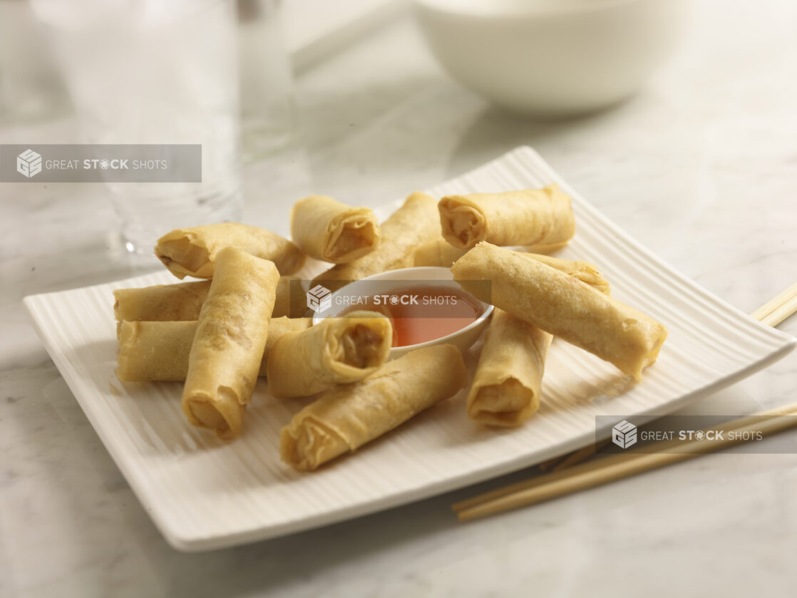 Deep fried spring rolls with plumb sauce on a white square platter with chopsticks on a white marble background