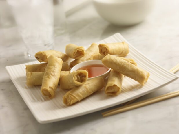 Deep fried spring rolls with plumb sauce on a white square platter with chopsticks on a white marble background