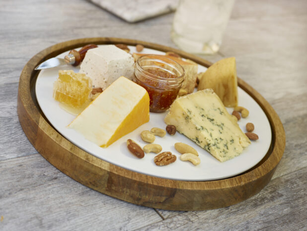 Round cheese tray with various cheeses, nuts, honeycomb and red pepper jelly in a small mason jar on a grey wooden background