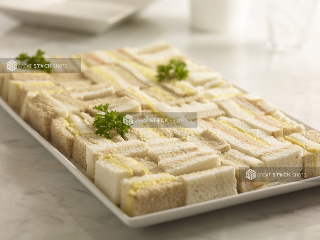 Assorted egg salad, tuna salad and salmon salad party sandwiches, on a white rectangular platter on a white marble background