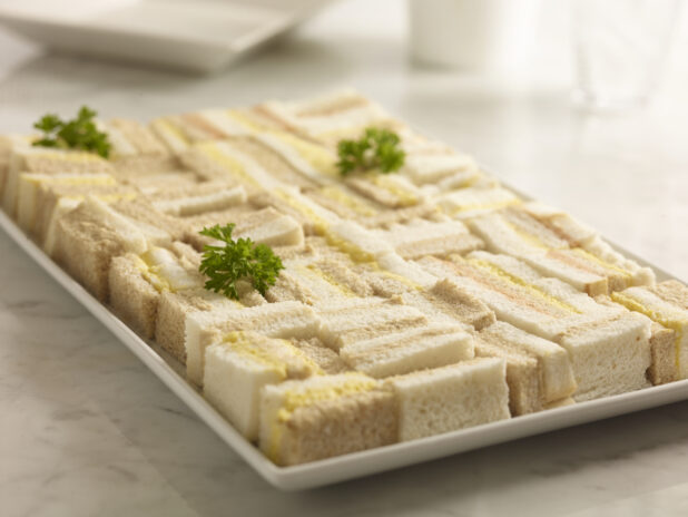 Assorted egg salad, tuna salad and salmon salad party sandwiches, on a white rectangular platter on a white marble background