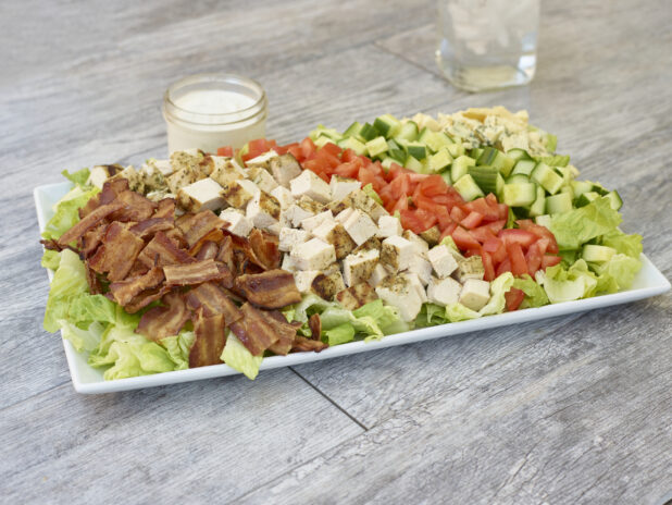 Chicken BLT salad with chicken, bacon, tomatoes, cucumber and blue cheese on a white rectangular platter with dressing in a mason jar on a grey wooden background