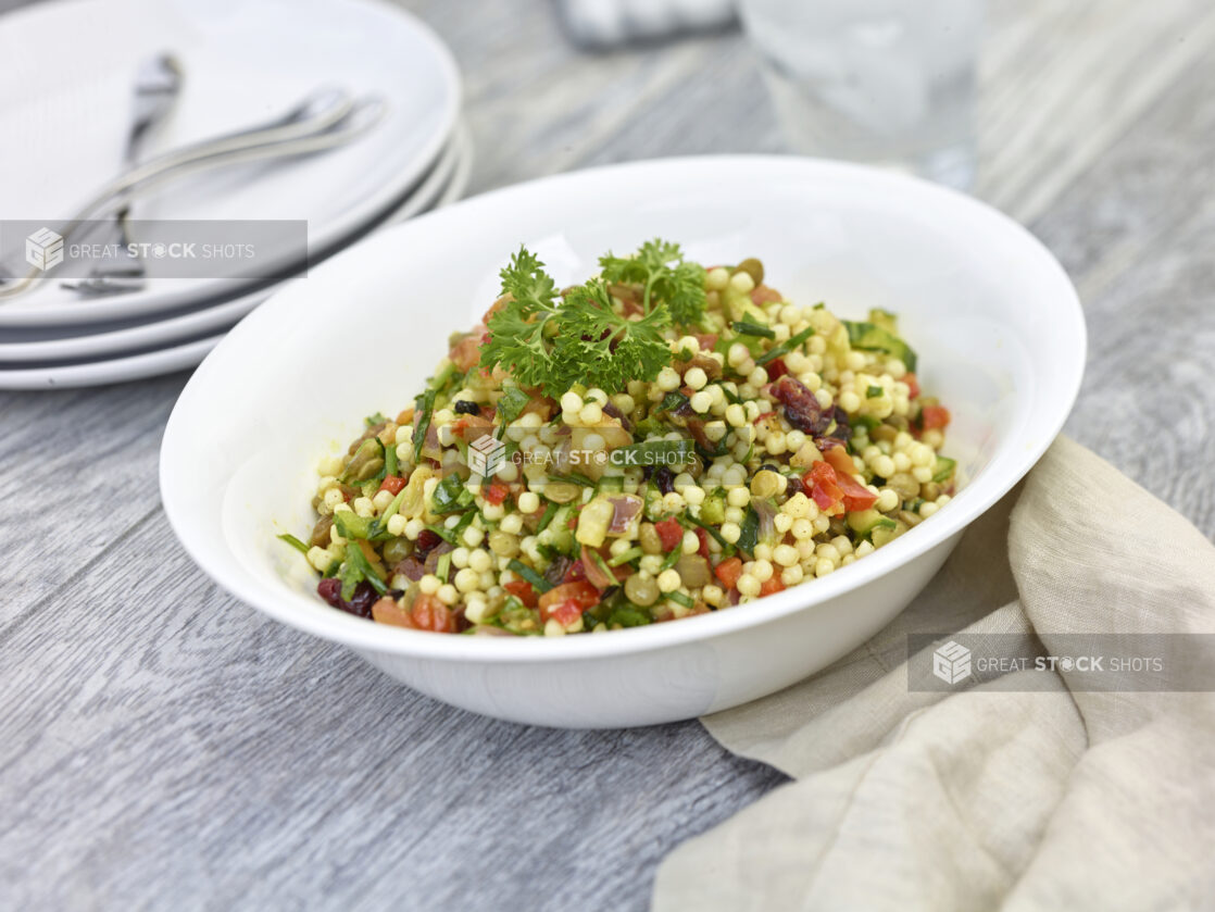 Fregola pasta salad in a round white bowl with plates and forks in the background on a grey wooden background