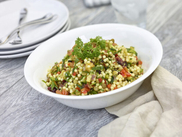 Fregola pasta salad in a round white bowl with plates and forks in the background on a grey wooden background
