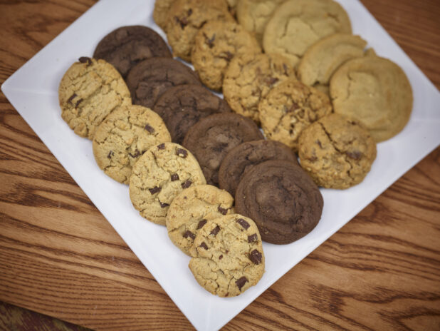 Assorted cookies, chocolate chips, double chocolate, oatmeal raisin and peanut butter on a square white platter on a wooden background