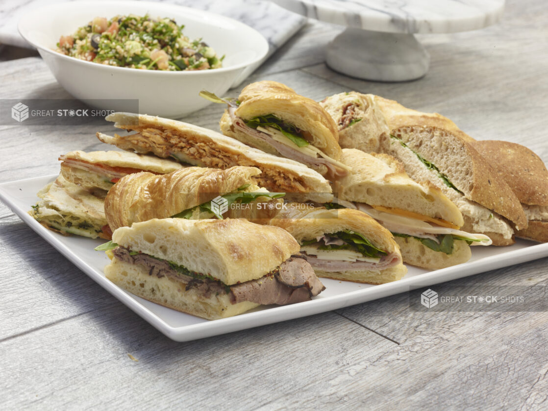 Assorted artisanal sandwich platter on a rectangular white platter with tabouli salad in a white bowl in the background on a grey wooden background