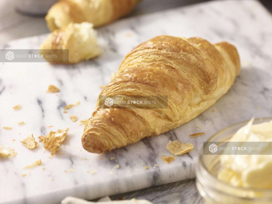 Croissant on a marble board with a torn croissant in the background a mason jar of butter in the foreground