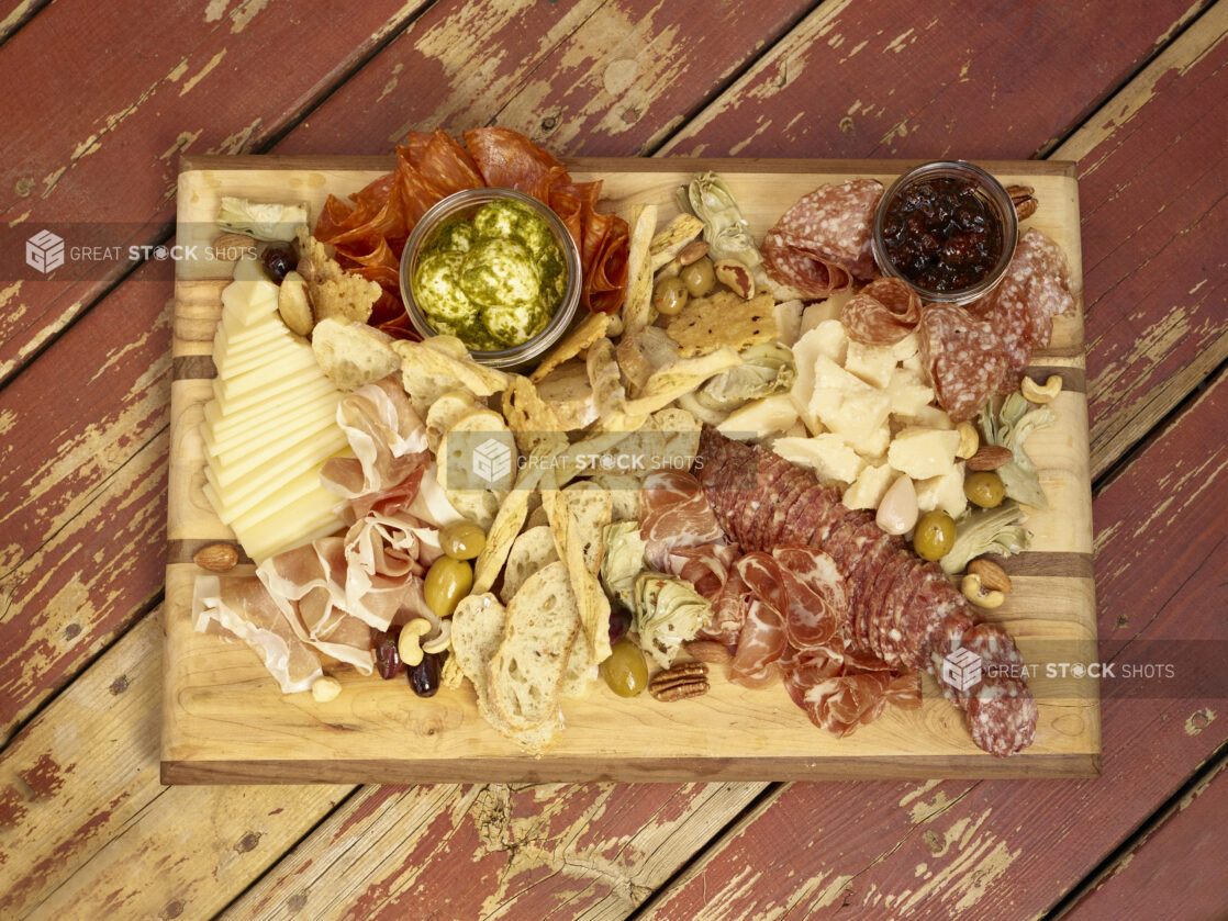 Charcuterie with salami, prosciutto, cheeses, olives, bread, crackers, nuts, jar of jelly, jar of pesto bocconcini on a wooden board on a rustic wooden background