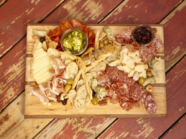Charcuterie with salami, prosciutto, cheeses, olives, bread, crackers, nuts, jar of jelly, jar of pesto bocconcini on a wooden board on a rustic wooden background