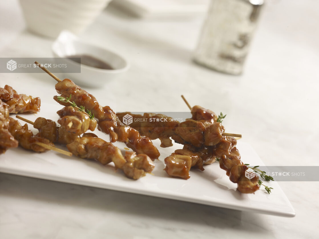 Chicken satay on a rectangular white platter with thyme sprigs with dipping sauce on the side on a white marble background