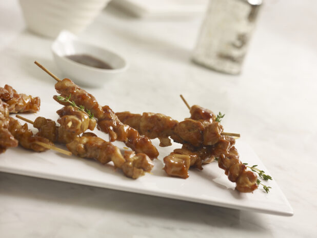 Chicken satay on a rectangular white platter with thyme sprigs with dipping sauce on the side on a white marble background