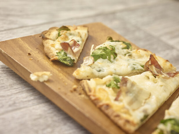 Cut flatbread with arugula, bacon and cheese in a close up view on a wooden board on a grey wooden background