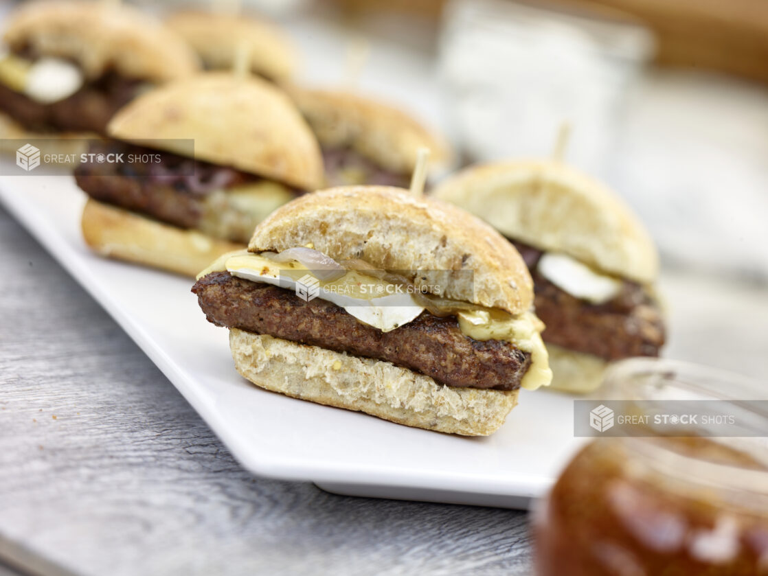 Hamburgers with brie cheese on ciabatta buns on a white rectangular platter, close up view, on a grey wooden background