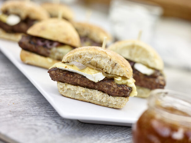 Hamburgers with brie cheese on ciabatta buns on a white rectangular platter, close up view, on a grey wooden background