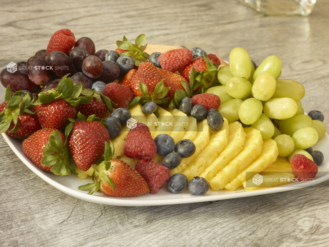 Fruit platter on a rounded square plate with pineapple, strawberries, grapes, blueberries, cantaloup on a wooden background