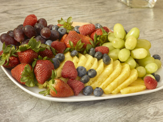 Fruit platter on a rounded square plate with pineapple, strawberries, grapes, blueberries, cantaloup on a wooden background
