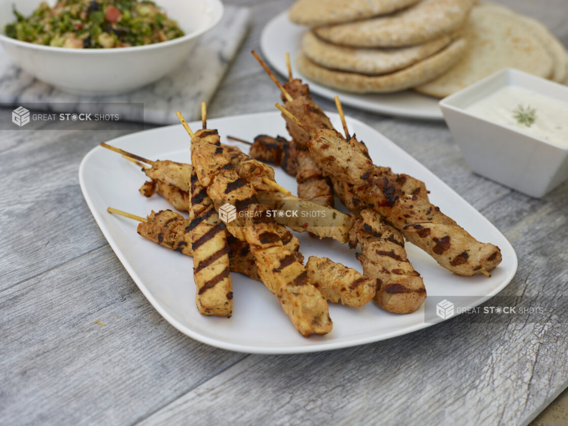 Chicken skewers on a rounded square plate with a healthy salad and mini pitas in the background on a grey wooden background