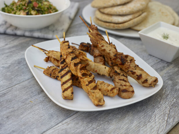 Chicken skewers on a rounded square plate with a healthy salad and mini pitas in the background on a grey wooden background