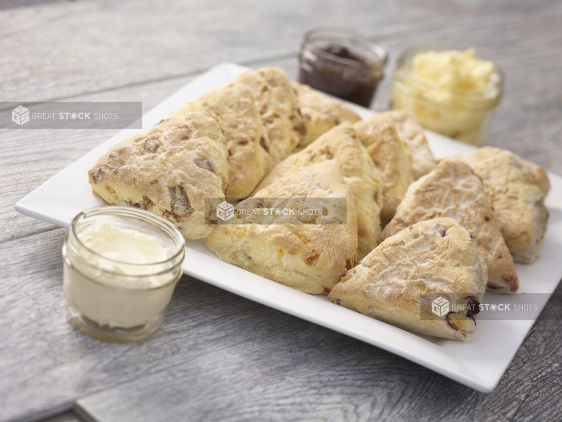 Assorted tea biscuits/scones on a white square platter with butter, jam and cream cheese in mason jars on a grey wooden background