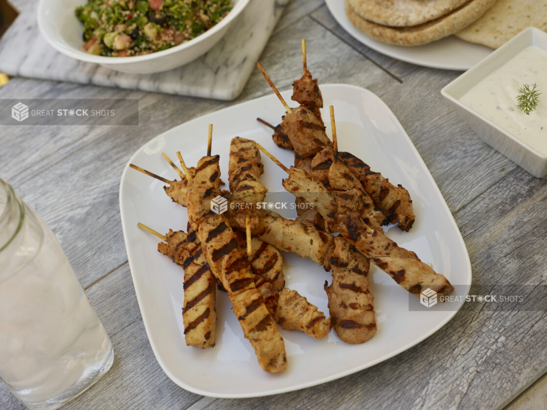 Middle Eastern chicken skewers on a square rounded plate with, salad, pitas and dipping sauce on the side on a grey wooden background