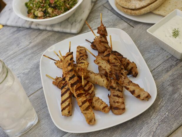 Middle Eastern chicken skewers on a square rounded plate with, salad, pitas and dipping sauce on the side on a grey wooden background